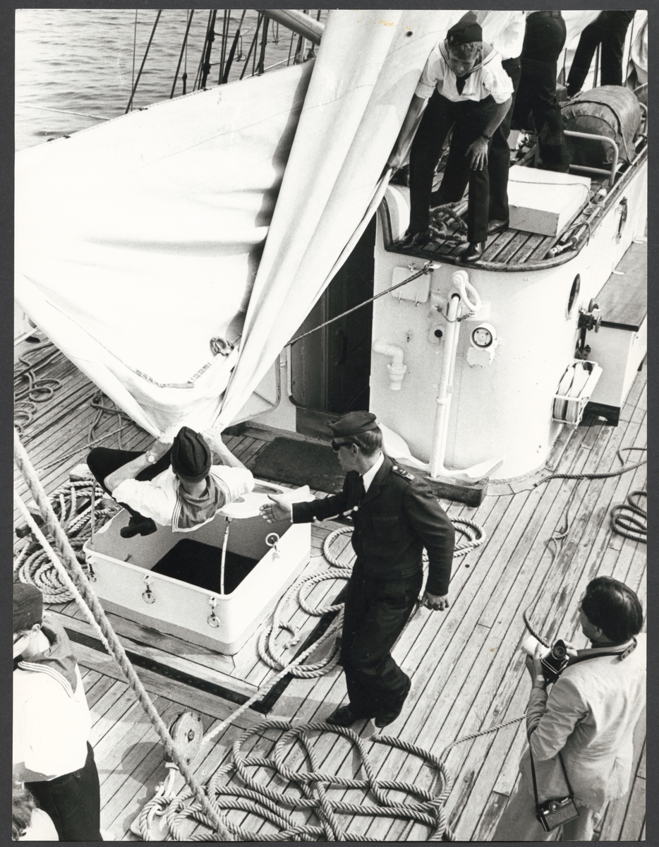 Bilden visar kadetter på en av Marinens skolfartyg som håller på att bärga ett segel. I mitten ser man en sjöofficerare som ge anvisningar. Längst ner i höger hörnan syns en pressfotograf som ta kort av sceneriet.