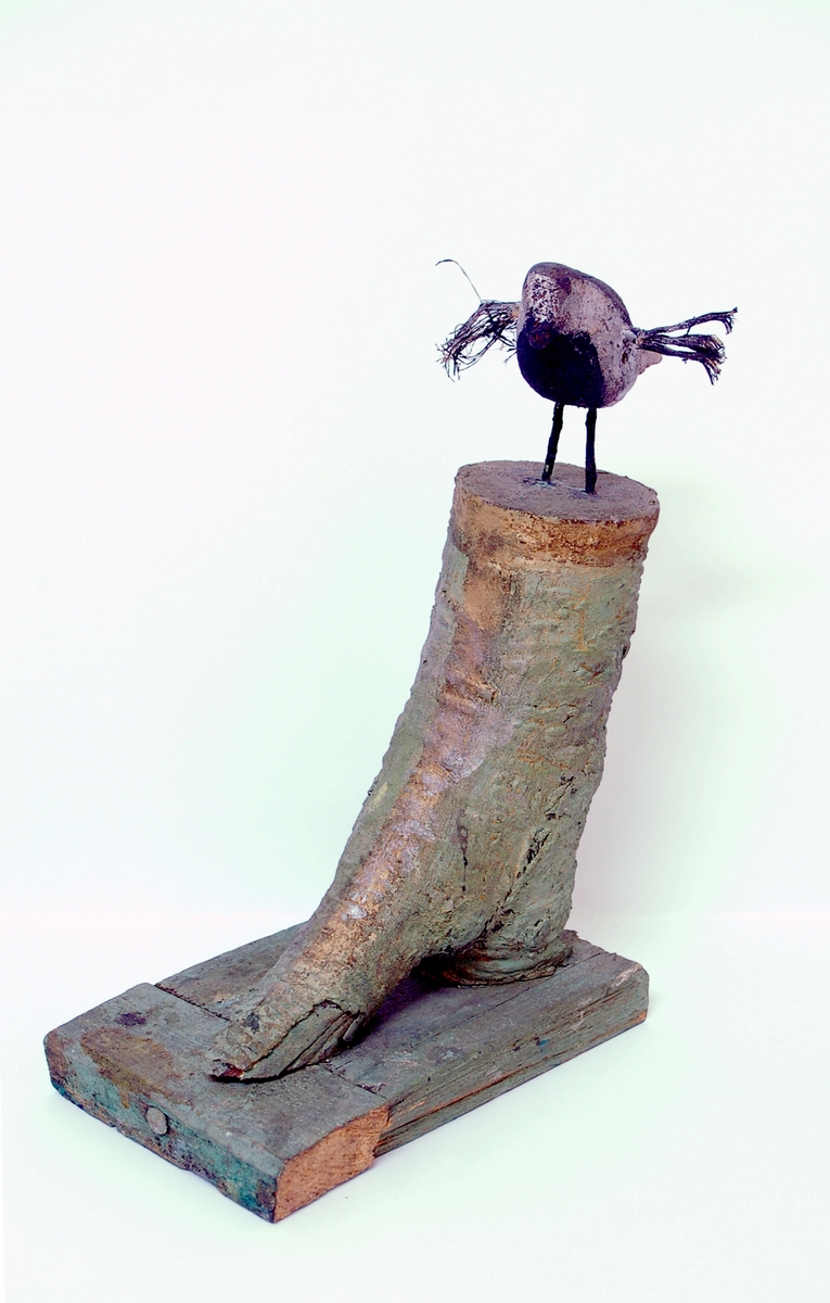 Skulptur av trä. Skulpterad/snidad fågel placerad på en fotliknande sockel, vilken är skapad av en grenklyka. "Foten" är grönmålad, fågeln målad randig i svart och silver, med utbredda vingar av (troligen) tagel.