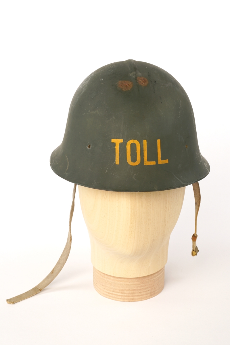 Metall hjelm (magnetisk), grønnmalt med 'TOLL'  stensilerte i gul maling. På innsiden, tre lær klaffer med puter på innsiden, forbundet med en sirkel av snor å justere for hodestørrelse. Tekstilbånd på hver side, en med metalllås for lukking under haken.