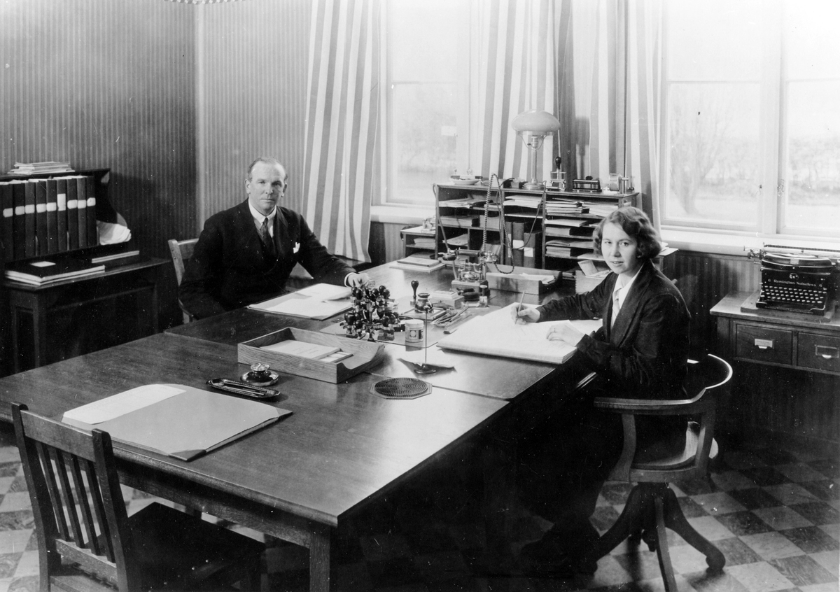 Kontorsrum med en man och en kvinna på var sin sida av ett större skrivbord. Inifrån Alingsås lasarett.