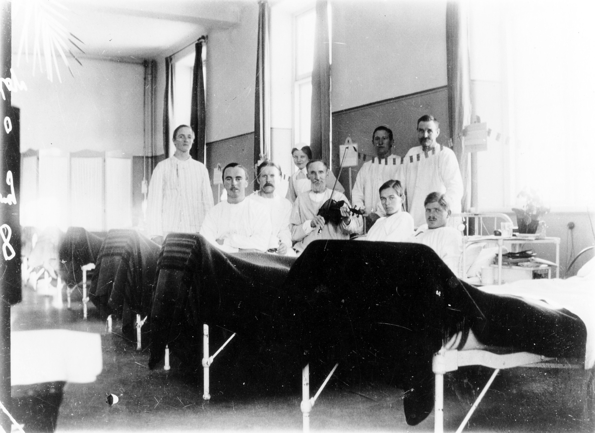 Bild tagen inifrån en sjuksal på Alingsås lasarett med 8 manliga patienter och en kvinnlig sjuksköterska. Mannen mad fiolen i mitten är Johannes Eriksson, pappa till fotografen Teodor Eriksson.