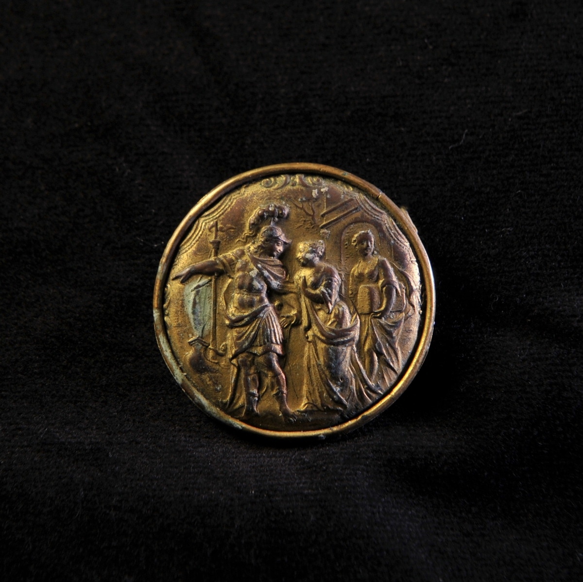 En rund brosch av bronserad mässing med antikt motiv i relief, i form av krigare med två kvinnor.