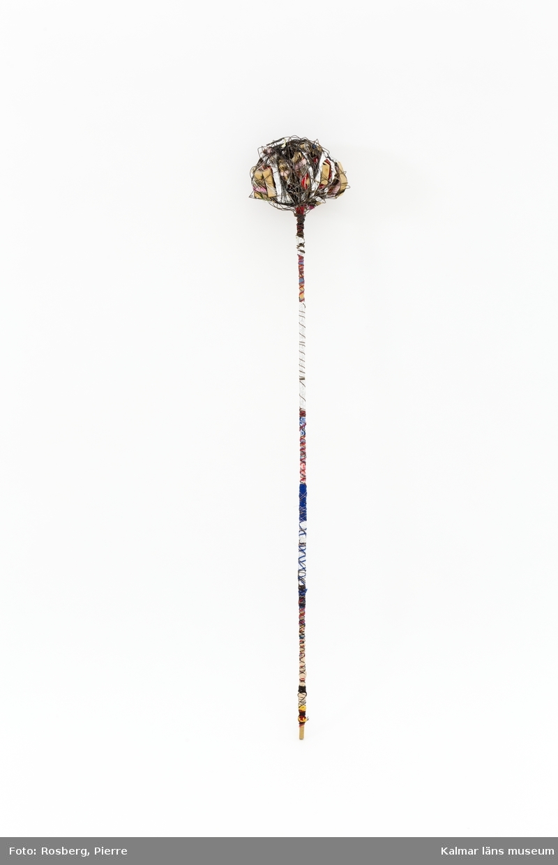 En stav av bambu lindad med band och garn. Högst upp ett klot av tändsticksasklådor som lindats ihop med metall- och sytråd.