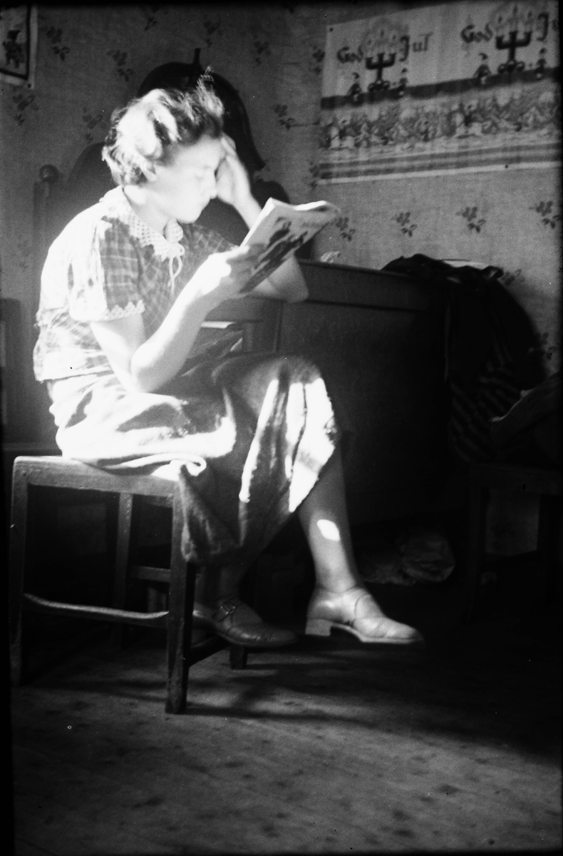 En flicka sitter vid fönstret och läser en tidning vid en julbonad med ljusstakar, tomtar och nedtill ridande män i granskog. Flickans blus har virkad krage och kant på ärmen.