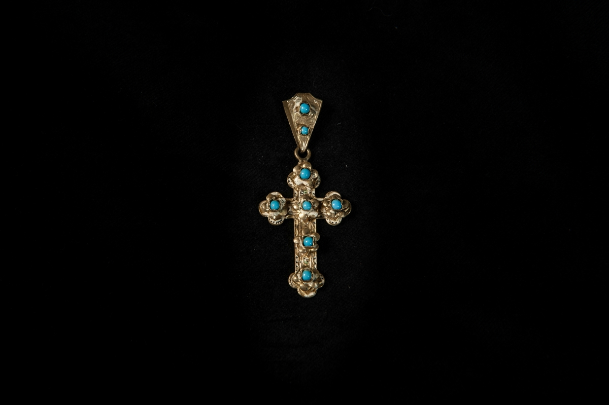 Ett hängsmycke i form av ett kors i gulmetall med gjuten dekor, med sex stycken infattade små turkoser på korset, samt två infattade turkoser på öglan. Är enligt givaren inköpt i Italien.