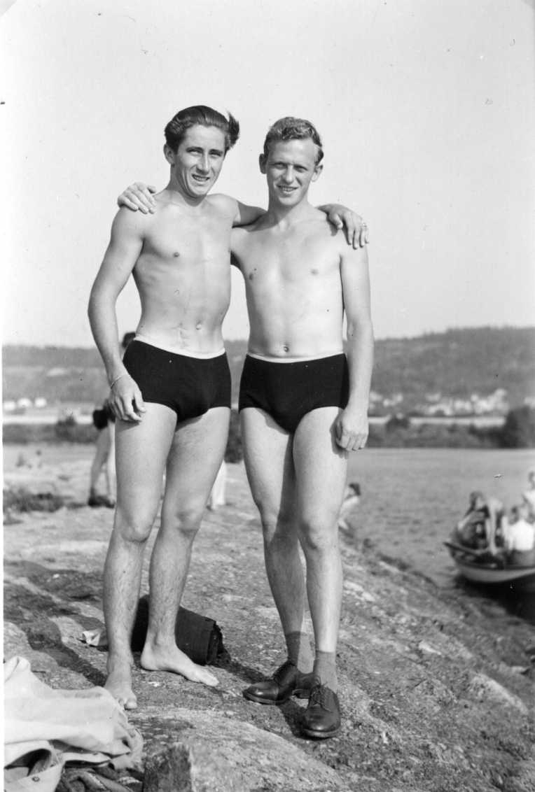 Två unga män i badbyxor med arm på varandras axel. Troligen på Visingsö med Gränna i bakgrunden.