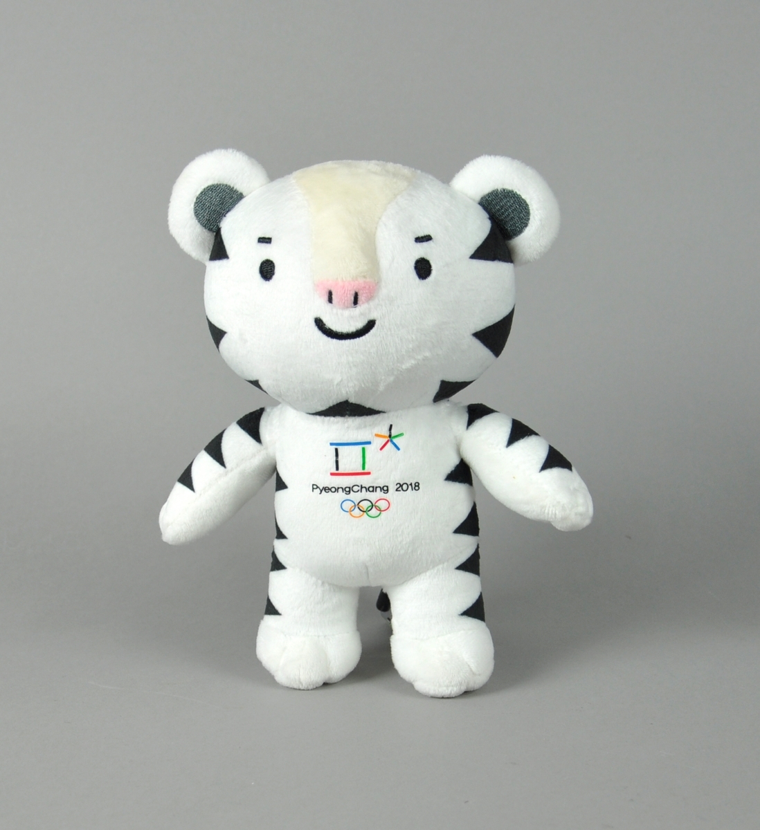 Hvit bamse med svarte flekker. På brystet vises logo for vinter-OL i PyeongChang 2018 med de olympiske ringene under.