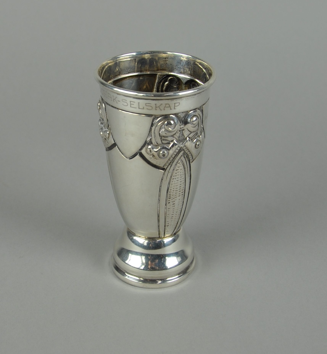 Pokal av sølv. Pokalen har sylindrisk form med innsnevret bunn. Utgående sokkel med profilert form. På korpus er det gravert dekor i dragestil.