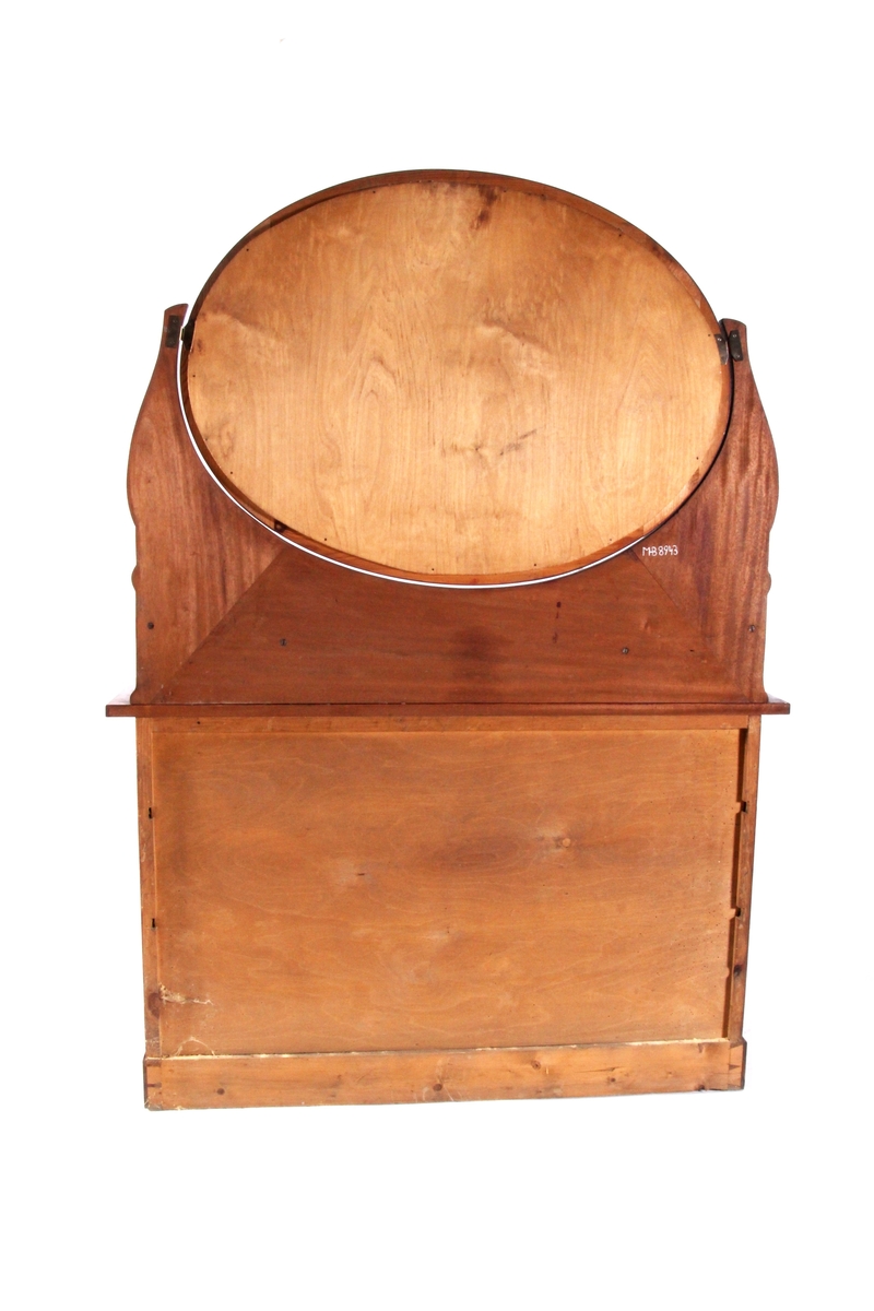 Brunlakkert toalettkommode med ovalformet speil som kan vippes fram og tilbake. Skuffeseksjon med to småskuffer på hver side av speilet. Underdelen består av tre langskuffer som er låsbare, med håndtak og nøkkelskilt