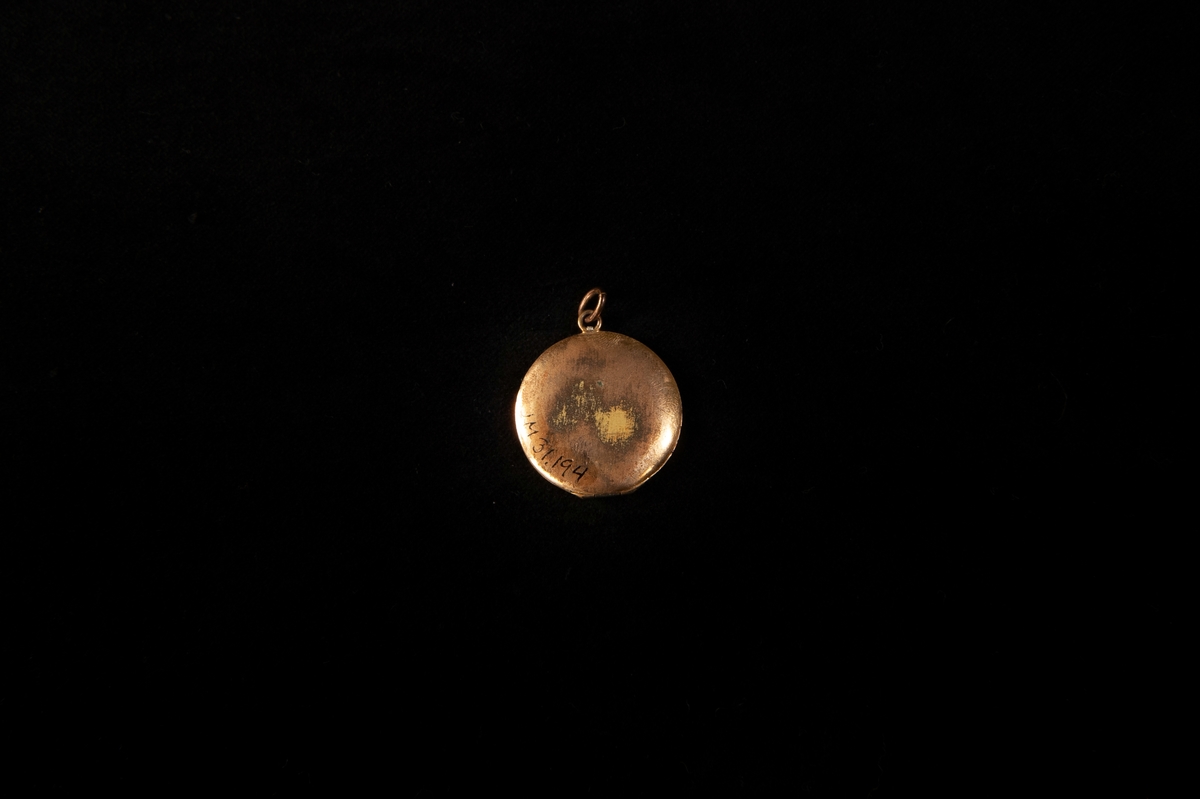 En rund medaljong i förgylld mässing. På framsidan finns ett graverat mönster och en infattad vit pärla. Inne medaljongen finns två fotografiier. Baksidan är sliten och där har den ärgat.