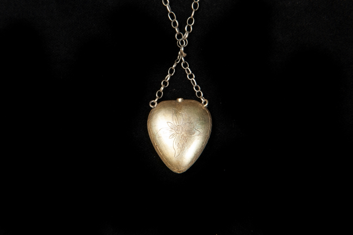 Enkel halskedja av metall med hänge i form av  ett ihåligt hjärta av silver. Graverad dekor i form av en blomma framtill. Baktill en zick-zackbård. Litet hål upptill. Hjärtat har varit avsett för så kallat luktvatten. Stämplad (något otydligt) bakpå.