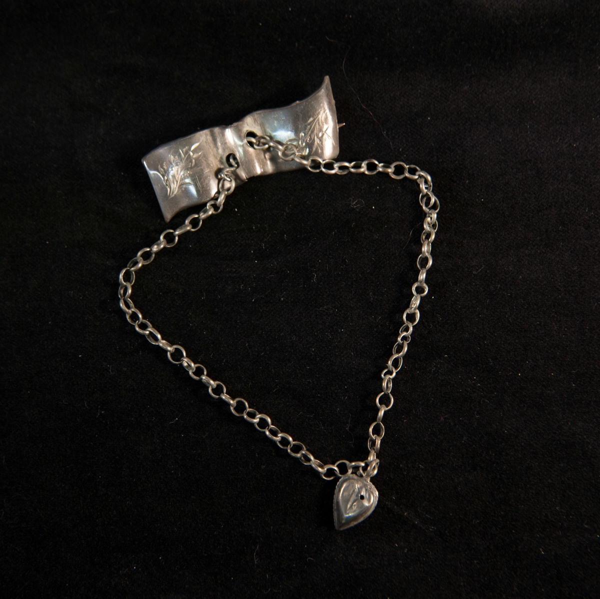 En rektangulär brosch av silverliknande metall med graverad bladdekor. I mitten hänger en kedja med ett pressat hjärta av samma metall. Saknar stämplar.