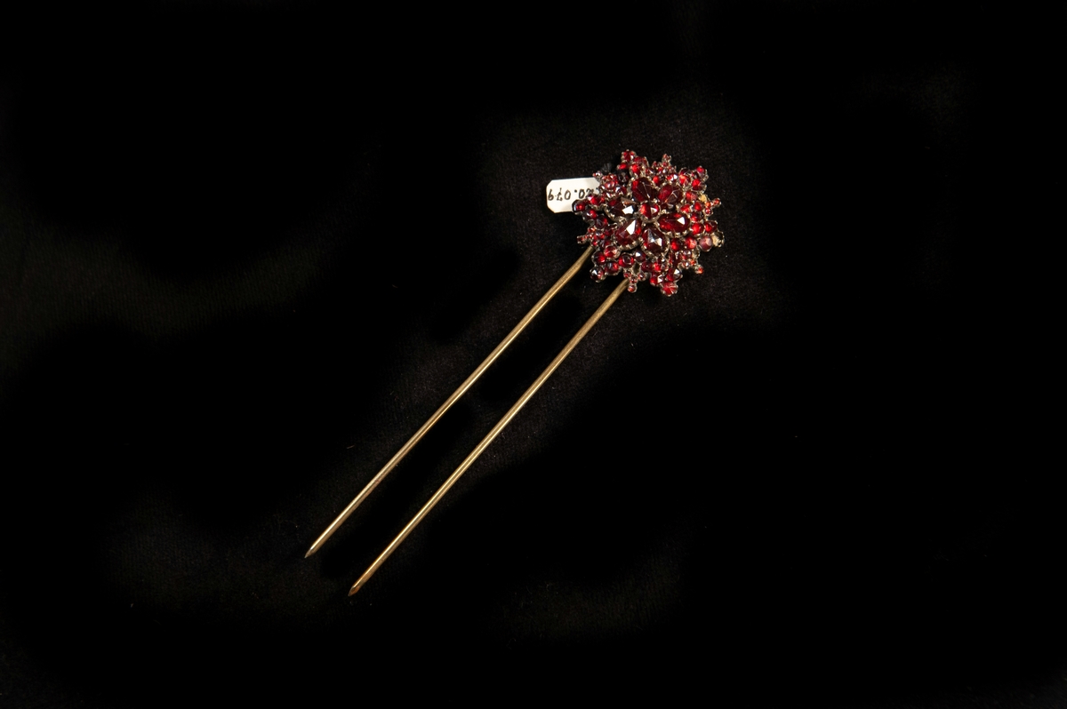 Hårnål av mässing i form av en sexuddig stjärna med infattade 
röda granater, delvis genombruten. Ledbar i toppen.