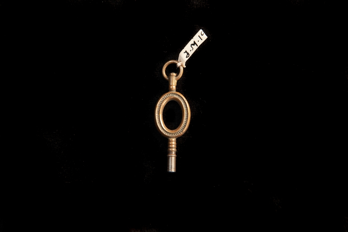 En urnyckel av brun metall (koppar?) med ett ovalt handtag med ett ovalt hål i, dekorerat med ringar längs kanten och streck däremellan.