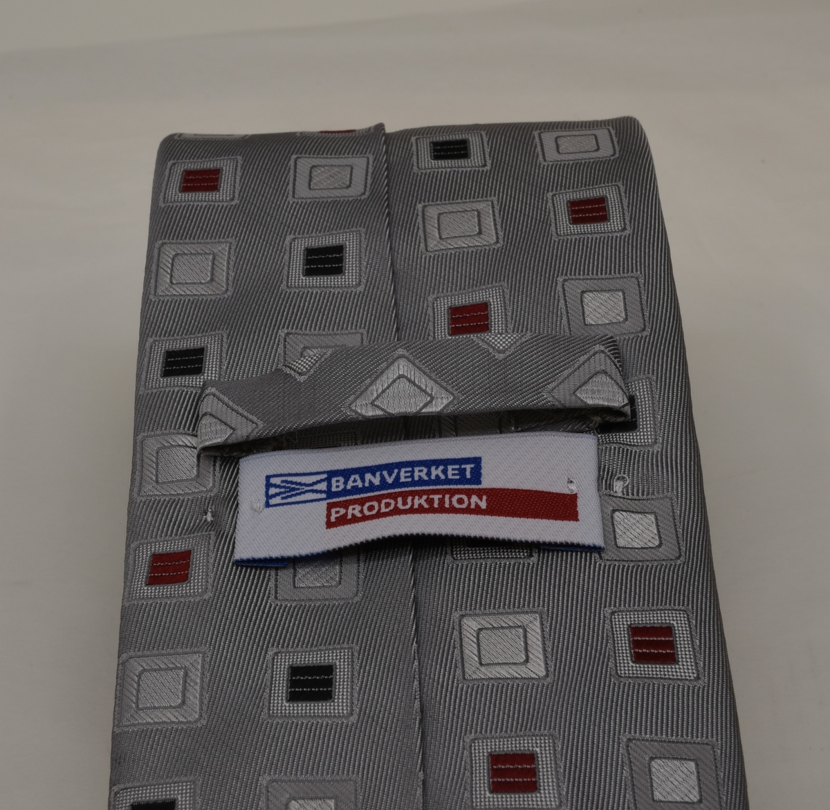 Grå slips av siden med upprepande mönster av kvadrater i färgerna svart, rött och grått. På baksidan av slipsen sitter två lappar.
