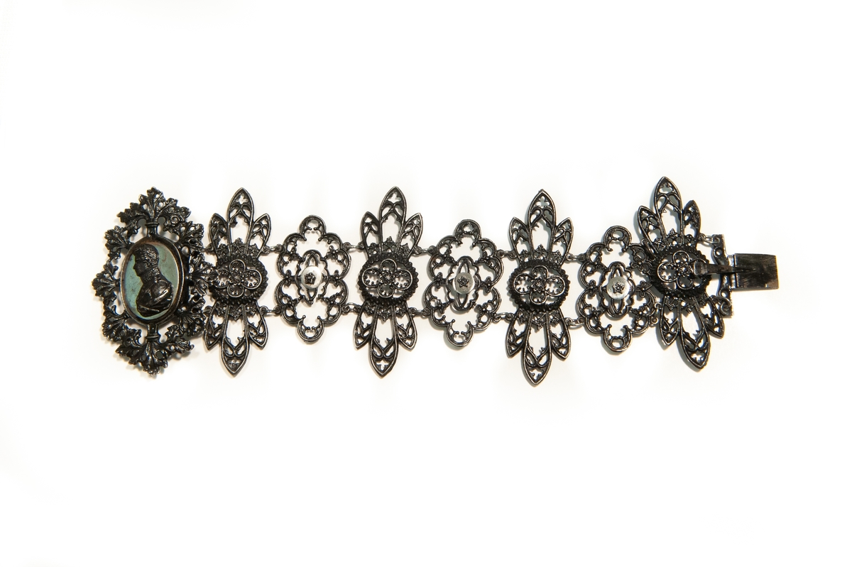 Armband av järn i åtta ledande delar, rikt genombrutna. På spännet som utgör en av delarna en bröstbild av man i profil på en spegelblank platta. Smycket är likt halsband JM 16.646:34.