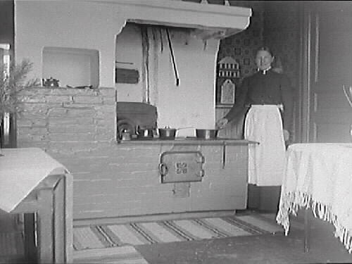 Spisgruvan i köket hos Maria Elmgren, kv Magistern, Prästgatan, med hennes jungfru stående vid spisen. Tallris och hyacint på borden antyder att bilden är tagen kring jul.