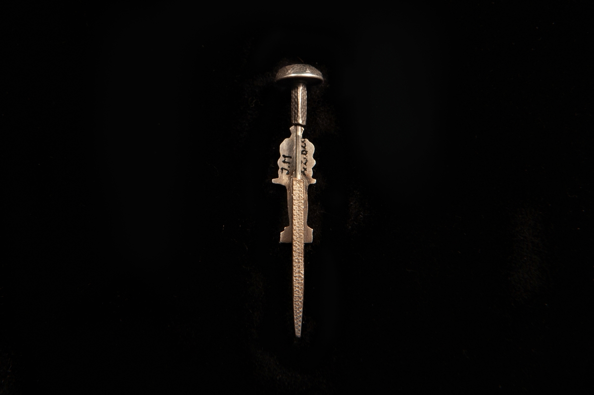 Kråsnål av metall i form av en stående riddare i rustning med svärdsbalja bakom. På nålens huvud ingraverat en riddare i rustning med årtal på var sida: "19 15".