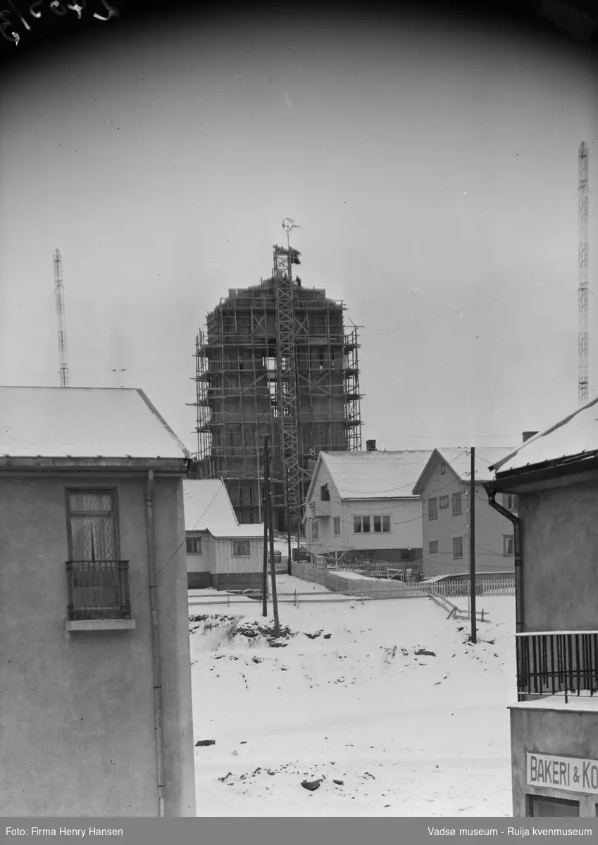 Vadsø 19.5.1956, kirka er under bygging. Kirketårnet har forskaling, og flagg og mønsåskrans på toppen. Bildet er tatt fra sentrum, foran til venstre litt av rådhuset, til høyre Henriksens bakeri.