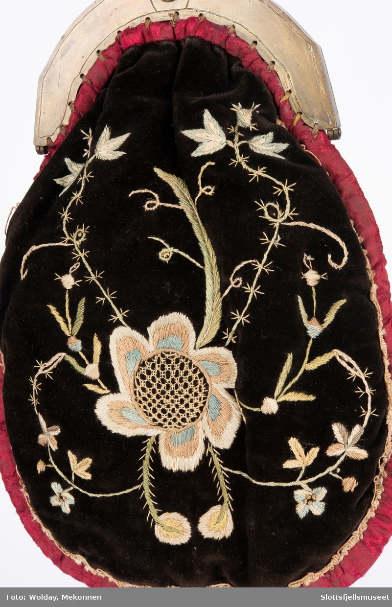 Pæreformet veske med bøyleformet lås. Brun fløyel er brodert med blomstermotiver. Rød kant rundt vesken.