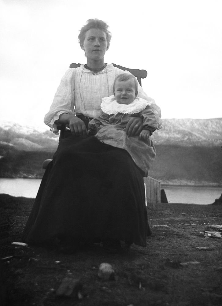 Leirfjord. Dame med ei lita jente, eller en gutt på fanget i en gyngestol, fotografert ute ved fjorden.