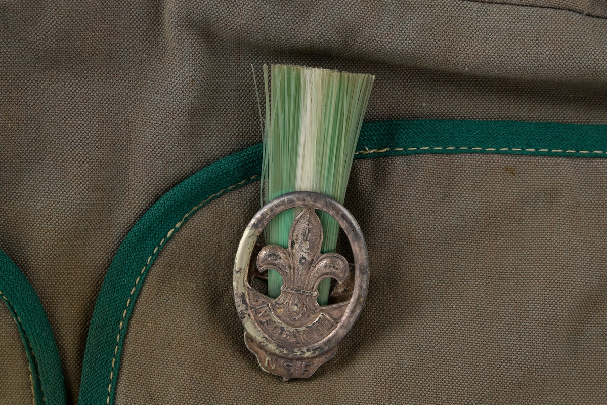 Tøymerke med speiderlilje i front. Metallnål på den ene siden av luen, med speiderlilje som motiv og farget bust.