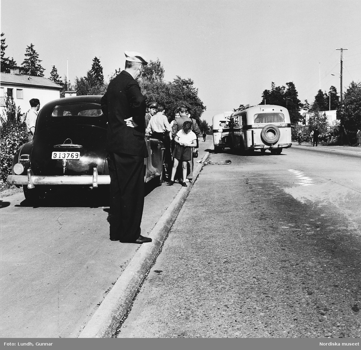 Två bussar har krockat på gamla Värmdövägen, Stockholm, år 1950. En man står på trottoarkanten och tittar, bakom honom en grupp människor vid en bil.