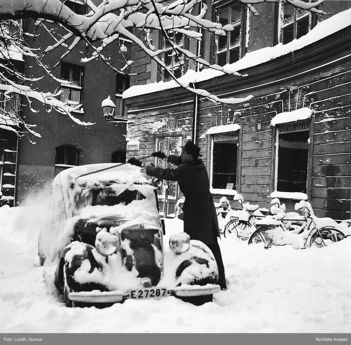 En man borstar av snön från en Mercedes 170, Gamla Stan, Stockholm år 1954. Marken är täckt av snö och vid husfasaden står snötäckta cyklar.