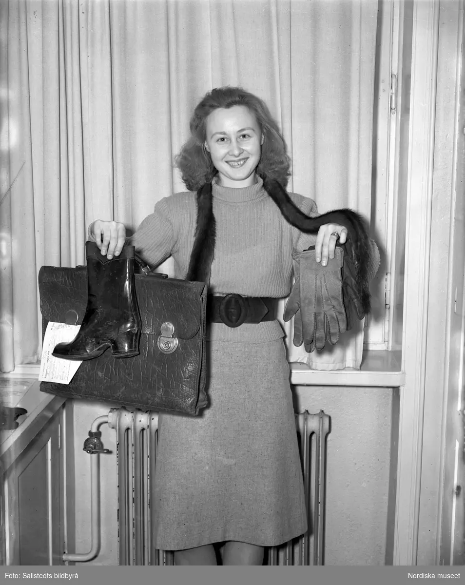 Enligt påskrift "Fru Marianne Ericsson på Solnapolisens hittegodsmagasin", år 1947. En kvinna håller upp inlämnat hittegods, handskar, portfölj och en stövel.