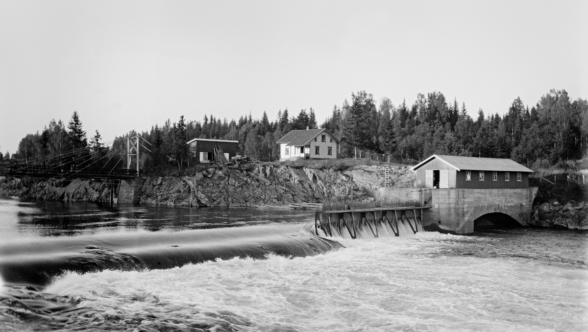 Bygging av nåledam - såkalt «fermettedam» - ved Svanfossen i elva Vorma i Fenstad i Nes kommune på Øvre Romerike, fotografert i 1916.  Den øvre delen av dette vassdraget ble noe regulert i slutten av 1850-åra, for å gjøre det mulig å kople den da nyetablerte hovedbanen med dampskipsruter på Mjøsa.  Den første dammen ved Svanfossen - den vi ser på dette fotografiet - skal ha vært bygd i 1910.  Her ser vi (til venstre i forgrunnen) hvordan elva faller over en terskel på elvebotnen.  På den østre delen av denne var det montert en såkalt «fermettedam», en stålkonstruksjon som bærer ei gangbru, der fløterne kunne gå når de skulle «sette dammen» (stenge) eller «sprette» (åpne) den. Dette ble gjort ved hjelp av «nåler», plank som ble stukket mot strømmen, slik at den lå an mot en terskel i botnen av elveløpet og mot gangbrua.  Disse nålene ble satt tett i tett, slik at de sammen dannet en vegg mot vannet.  Når dammen ble åpnet, brukte karene små kjetting- eller taustubber med ei jernnål i enden, som ble tredd gjennom hull i den øvre delen av nålene, og trakk til.  Ved siden av damkonstruksjonen ser vi et rektangulært hus med saltak på en høy mur, med en hvelvet åpning mot elveløpet under.  Dette kan muligens ha vært et lite kraftverk.  På bakkekammen sentralt i bildet lå det et  halvannenetasjes våningshus med kvite ytterveger, og ved siden av dette en rødmalt uthusbygning med saltak.