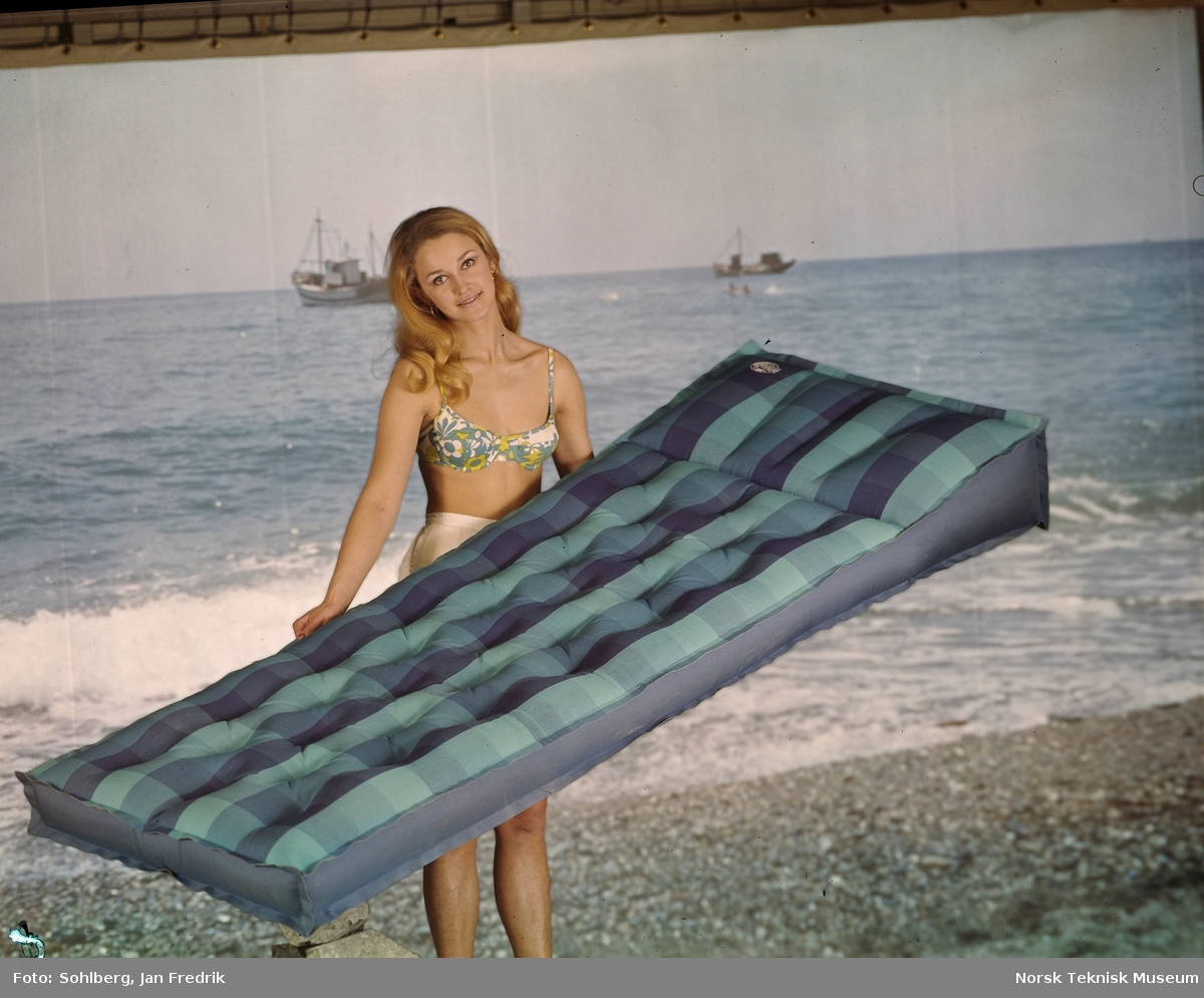En kvinnelig modell viser fra men bademadrass. Hun er forografert i studio mot en bakgrunn som forestiller hav og strand.