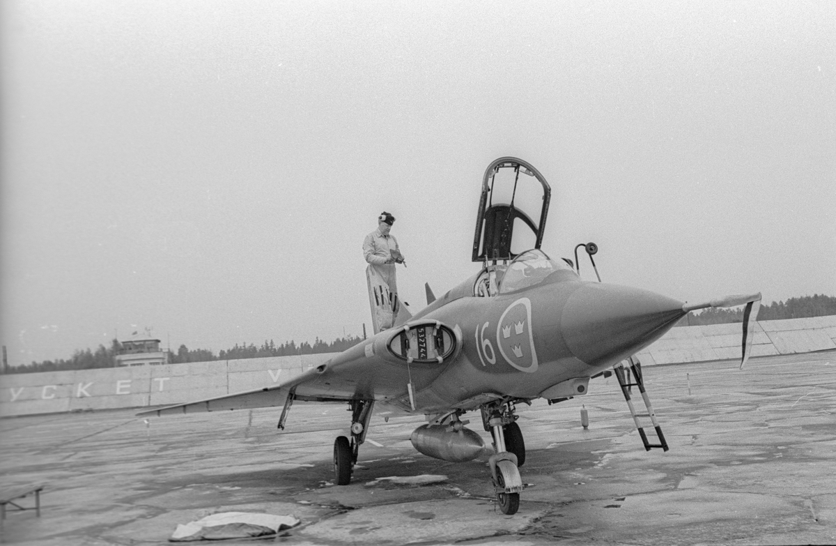 Flygplan J 35 Draken uppställd på sovjetisk flygbas. En man står på flygplanet. I bakgrunden skymtar texten 'Mycket välkomna' på en vägg. Vid Kungliga Upplands flygflottiljs besök vid sovjetisk MiG-21-bas den 16 mars 1972.