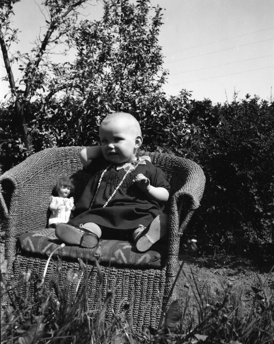 Ett litet barn med långt halsband sitter utomhus i en korgstol med en docka.
(Se även bildnr EA0891)