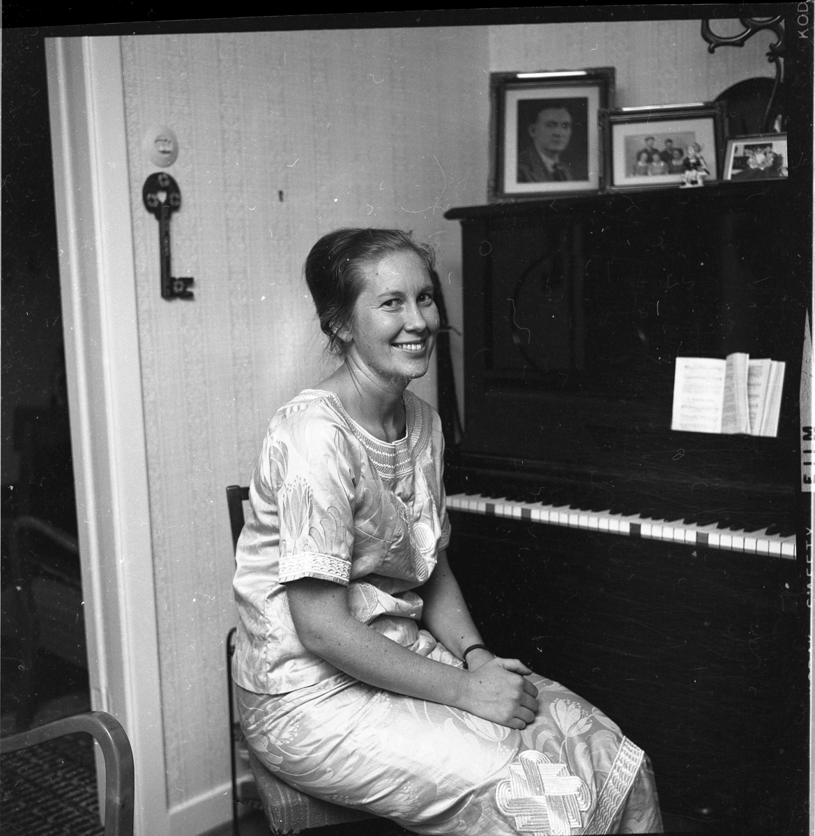 Marianne Boo sitter vid ett piano iförd en liberiansk festdräkt, en mönstrad klänning. På pianot står fotografier och på väggen till vänster hänger en stor nyckel.