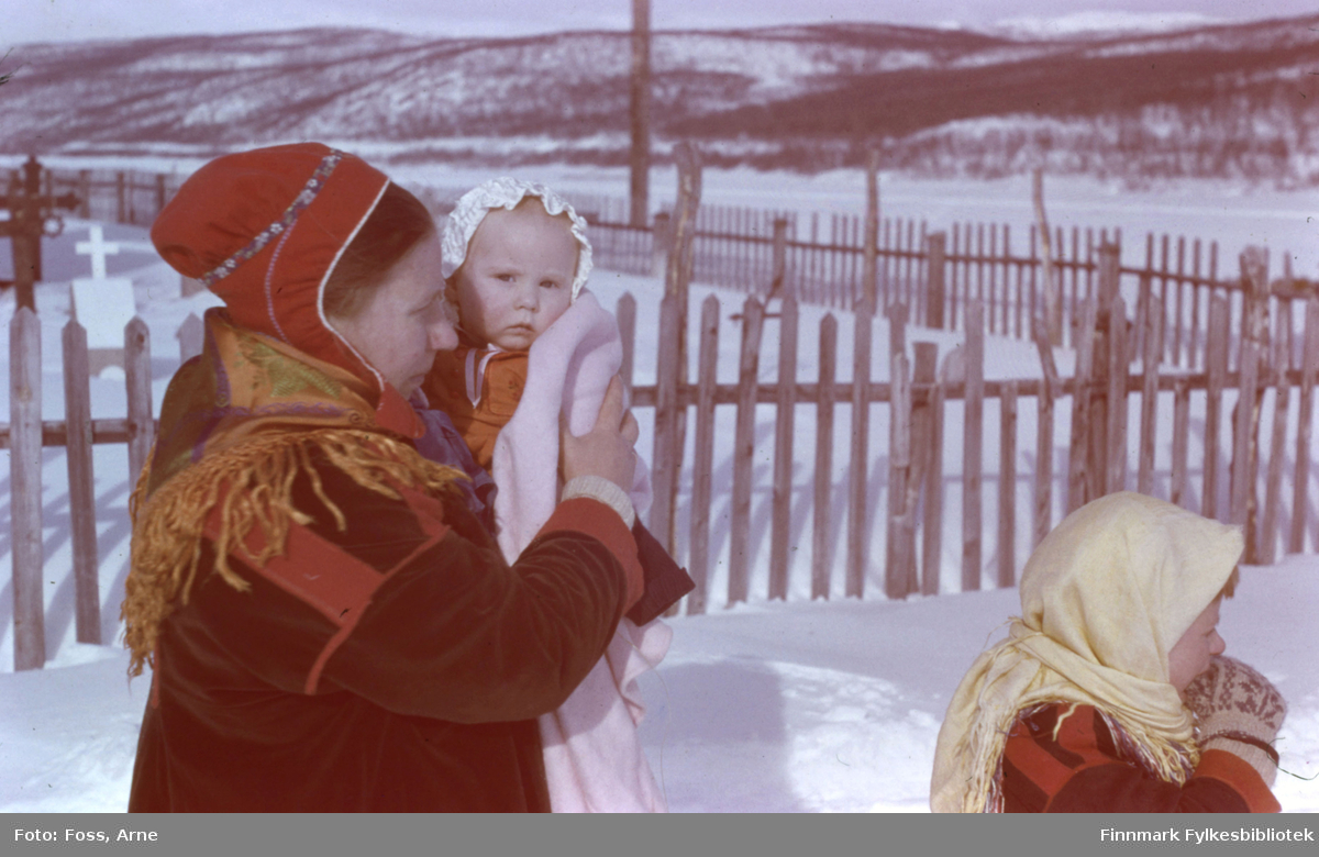 Den stolte mor Karen Saua (gift Eilertsen) holder sin datter Gunhild Sofie i armene til dåpen utenfor Polmak kirke, 2. påskedag i 1947. Ved siden står ei samejente som har hvitt skjerf rundt hodet. 