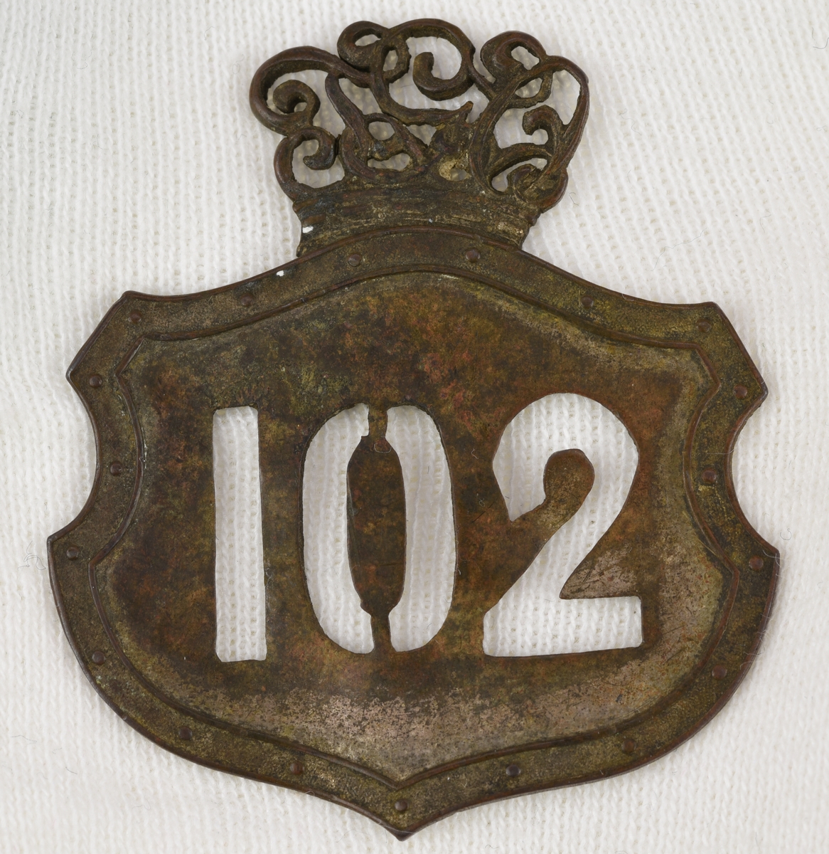 Stansad banvaktsplåt av mässing i form av en krönt sköld med en prickad bård. Siffrorna "102" är utstansade och kronan bildas av "TGO" i rikt ornerade bokstäver. Kronan är fastlödd mot märkets baksida. På baksidan syns spår av avbrutna fästnålar.