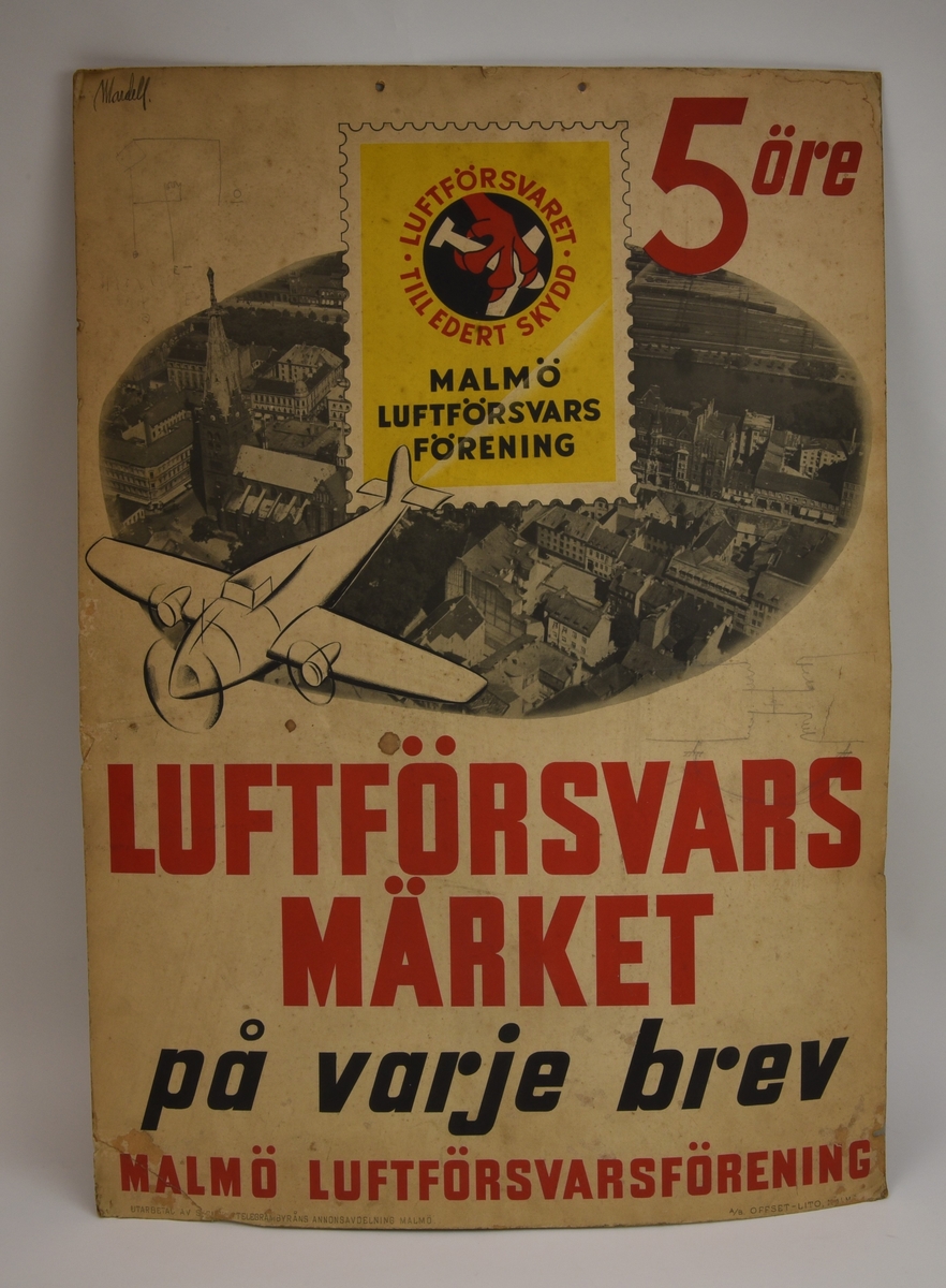 En reklamplansch, troligen 1930-tal, som gör reklam för Luftförsvarsmärket i Malmö.

Planschen har en ljusbrun bottenfärg. På planschens överdel finns en grafisk bild av ett gult frimärke med en röd gripklo som greppar ett vitt flygplan och som gör reklam för frimärken som stödjer Malmö Luftförsvarsförening. Till höger om frimärket finns ett pris angett med röd text "5 öre". På mitten av planschen finns ett flygfoto över Malmö och över det en grafisk bild av ett flygplan som flyger över staden. Planschens nedre halva är fylld med text: "

"LUFTFÖRSVARSMÄRKET
på varje brev
MALMÖ LUFTFÖRSVARSFÖRENING"

På planschens mederdel finns namnen på tryckeriet och beställaren (förmodligen).

Planschen verkar ha blivit återanvänd efter att den spelat ut sin roll som reklam för Malmö Luftförsvarsförening. I det övre vänstra hörnet finns ett efternamn skrivet med en blyertspenna: "Wandell". På planschens framsida finns även en teckning av ett elschema (?) samt en teckning av en transformator (?).

Planschens baksida har i efterhand återanvänts som plansch. På baksidan finns tre stycken olika förbindningsscheman/principscheman påklistrade som är märkta med "Telefon A.B. L.M. Ericsson".  Dessa tre olika scheman är ihoplänkade med varandra genom att någon med en blyertspenna har dragit linjer mellan olika punkter i schemorna, troligen för att knyta samman dem till ett större schema.