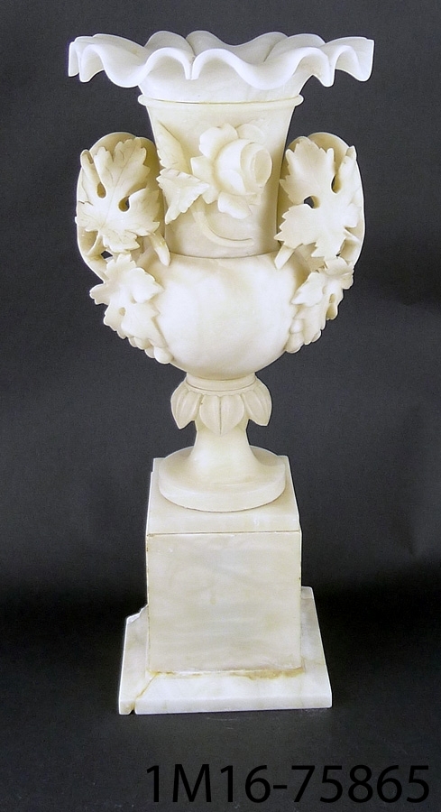 Urna av alabaster, fyrsidig sockel, på den en vas med vinrankor som öron och en ros på framsidan, veckad överkant.