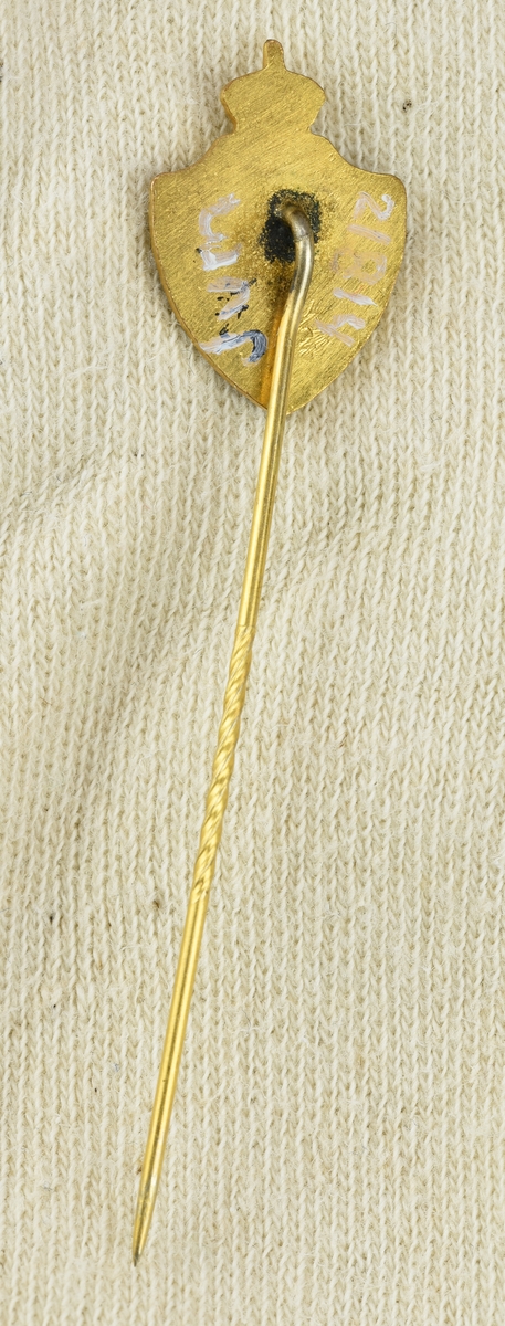 Kavajnål bestående av en platta i form av en sköld, krönt av ett bevingat hjul med krona, med en fastlödd nål på baksidan. Nålen är tillverkad av mässing med emalj. 
Skölden är indelat i tre fält i färgerna rött, gult och grönt.