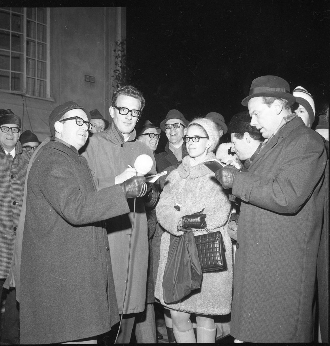 En grupp män och kvinnor, varmt klädda, är vända mot en okänd man i hatt till vänster som håller en mikrofon i handen. Närmast honom står Sten Sarborn med papper och penna i handen. Till höger står Björn Arding och antecknar. Till vänster skymtar Tage Grennfelt.