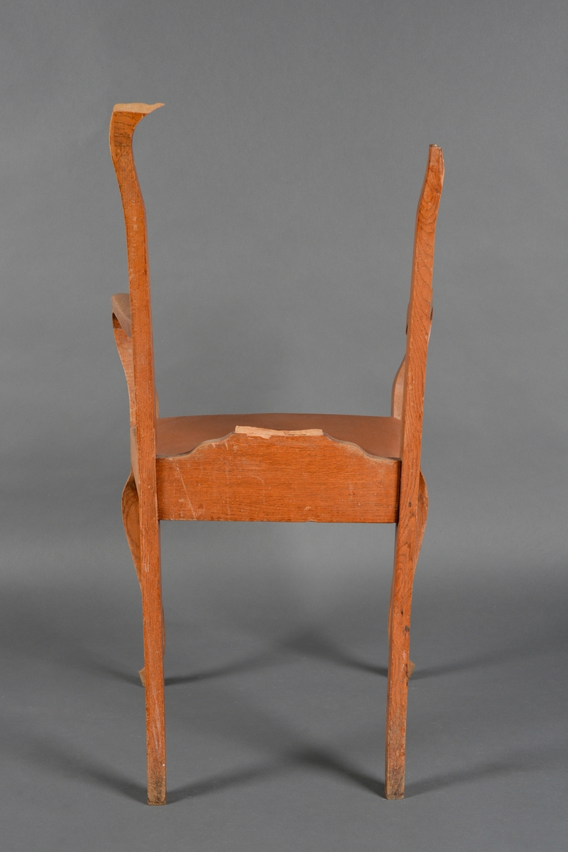 En stol laget av heltre med treforbindelser. Stolen har armelner som er buet i front. Ryggen er relativt høy og består av en bue med en midtplanke med enkle utskjæringer/dekor. Ryggen er noe ødelagt (toppen på buen og midtplanken er av). I setet er det skumgummi trukket med rød skai/skinnimitasjon. Treverket er lakkkert med blank lakk.