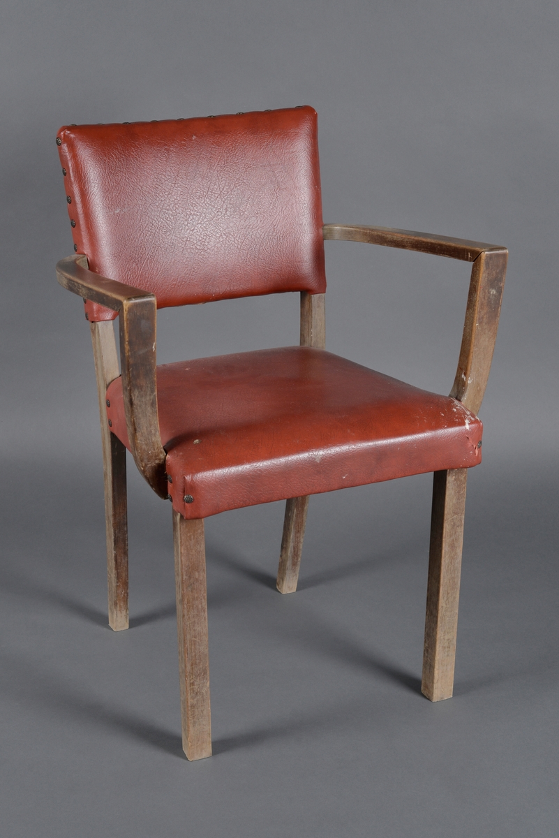 En stol med armlener laget av heltre og kryssfinér. Den har sammenføyninger med skruer samt treforbindelser. Treverket på stolen er heltre bortsett fra setet som er gjort i kryssfinér. Både rygg og sete er firkantet på form og er trukket med rød skai/skinnimitasjon. Trekket er festet med nagler som har stort rundt hode. Under dette trekket er det et tekstiltrekk som er originalt. Rygg og sete er polstret. Armlener og bein er firkantete på form. Treverket er beiset og lakkert.