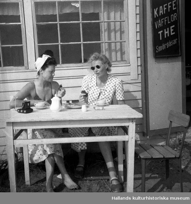 Två sommarklädda kvinnor dricker kaffe på en uteservering i Varbergstrakten. Till höger syns en skylt med texten "Kaffe Våfflor The Smörgåsar". På bordet ligger en kamera. Fotograf okänd.
