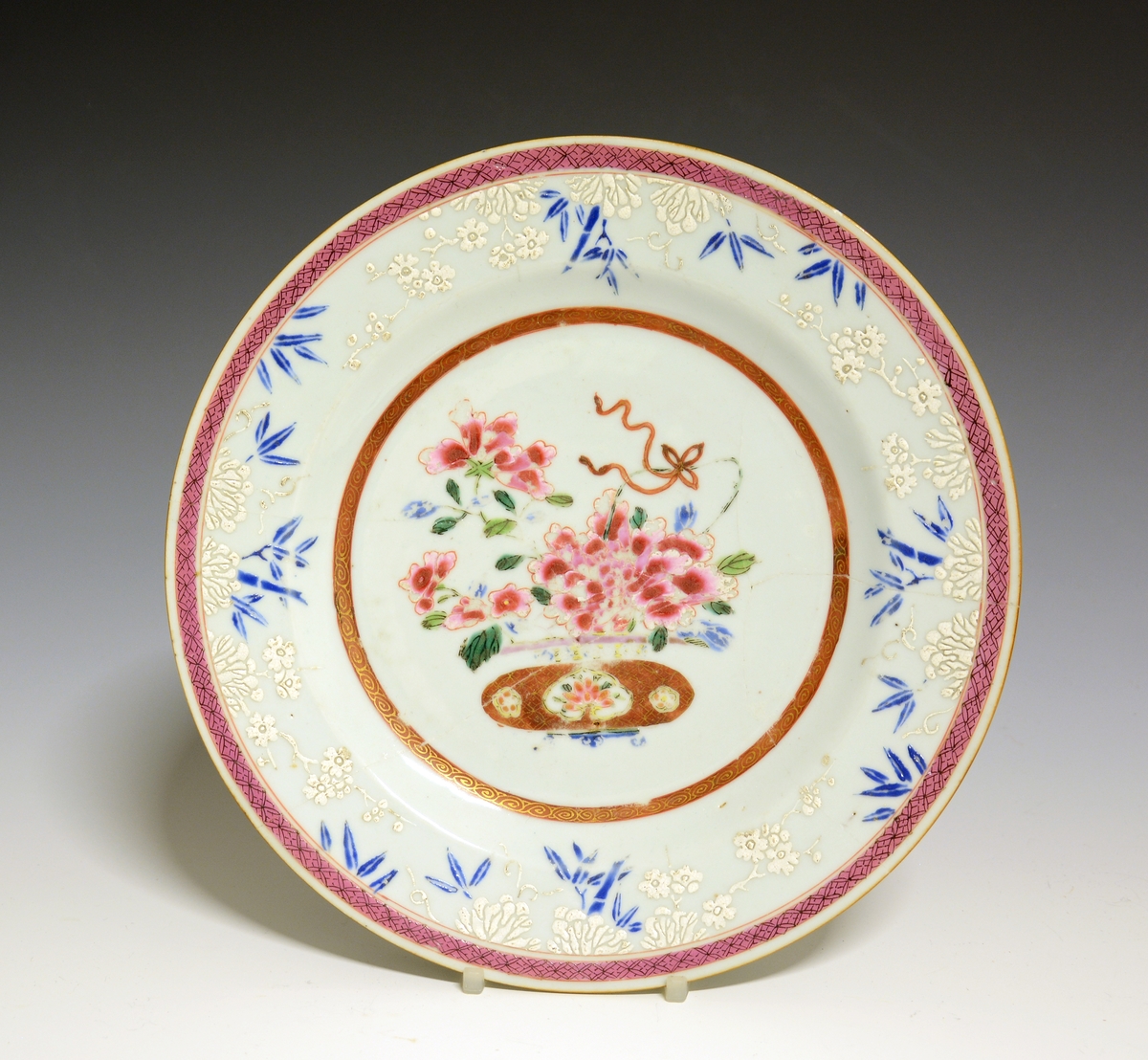 Flat, liten tallerken av porselen. Dekorert med blomster i en brun, lav blomstervase, vasen er dekorert med rosa blomster. Kina 1700 tallet. Famille rose. Langs fanen hvite og blå blomster. Det hvite ligger så tykt på at det danner et lavt relieff. Ytterst en rosa bord.