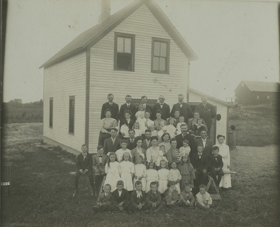 Ein gruppe vaksne og barn er oppstilt for fotografering, med eit lyst hus i bakgrunnen.