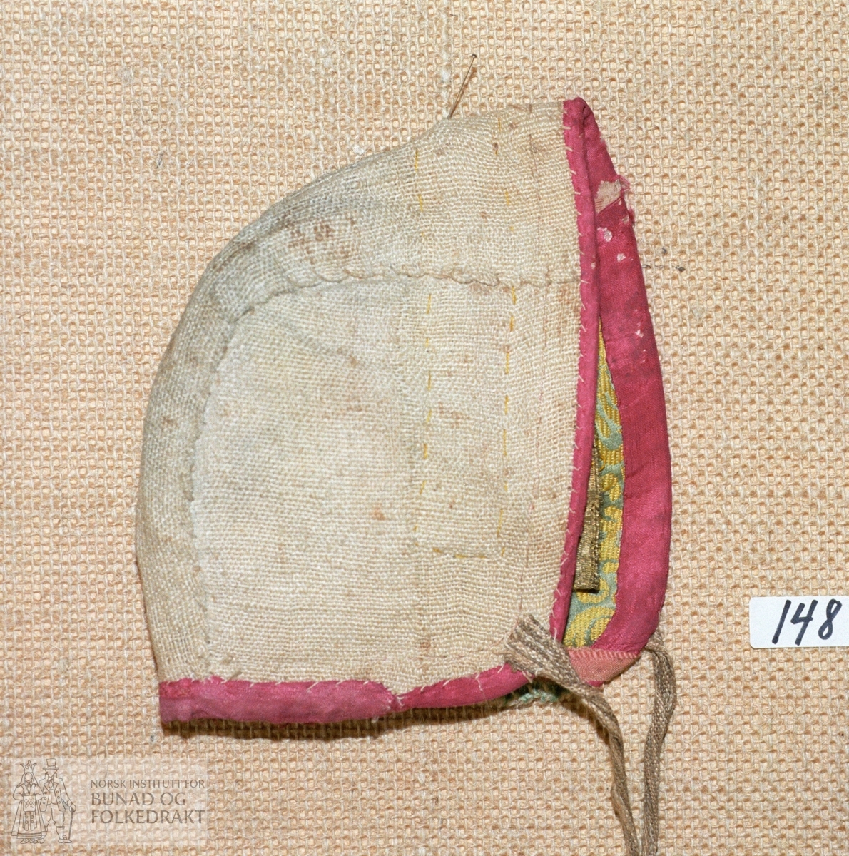 "Brukt i 1831".  Tobladslue laga av mønstra silkedamask fôra med linlerret. Kanta med blåraud silke. Prydd med gullband. Knyteband av stripa lin i ufarga og gulkvitt.
