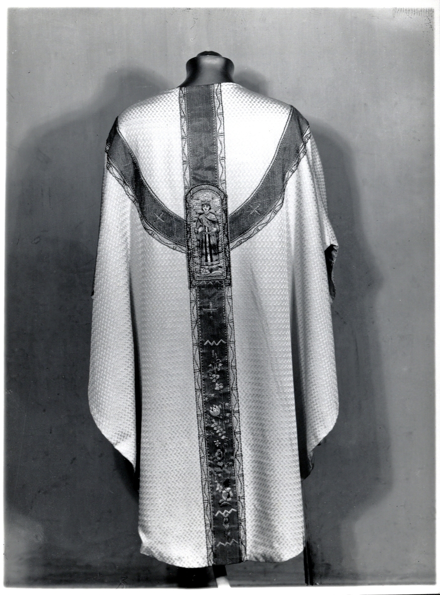 Foto (svart/vitt) av en ljus mässhake (baksidan) i brokad (?), med mörkare, broderat korsparti, prytt med bl a helgonbild, liksidiga kors och blommor.

Inskrivet i huvudbok 1983.