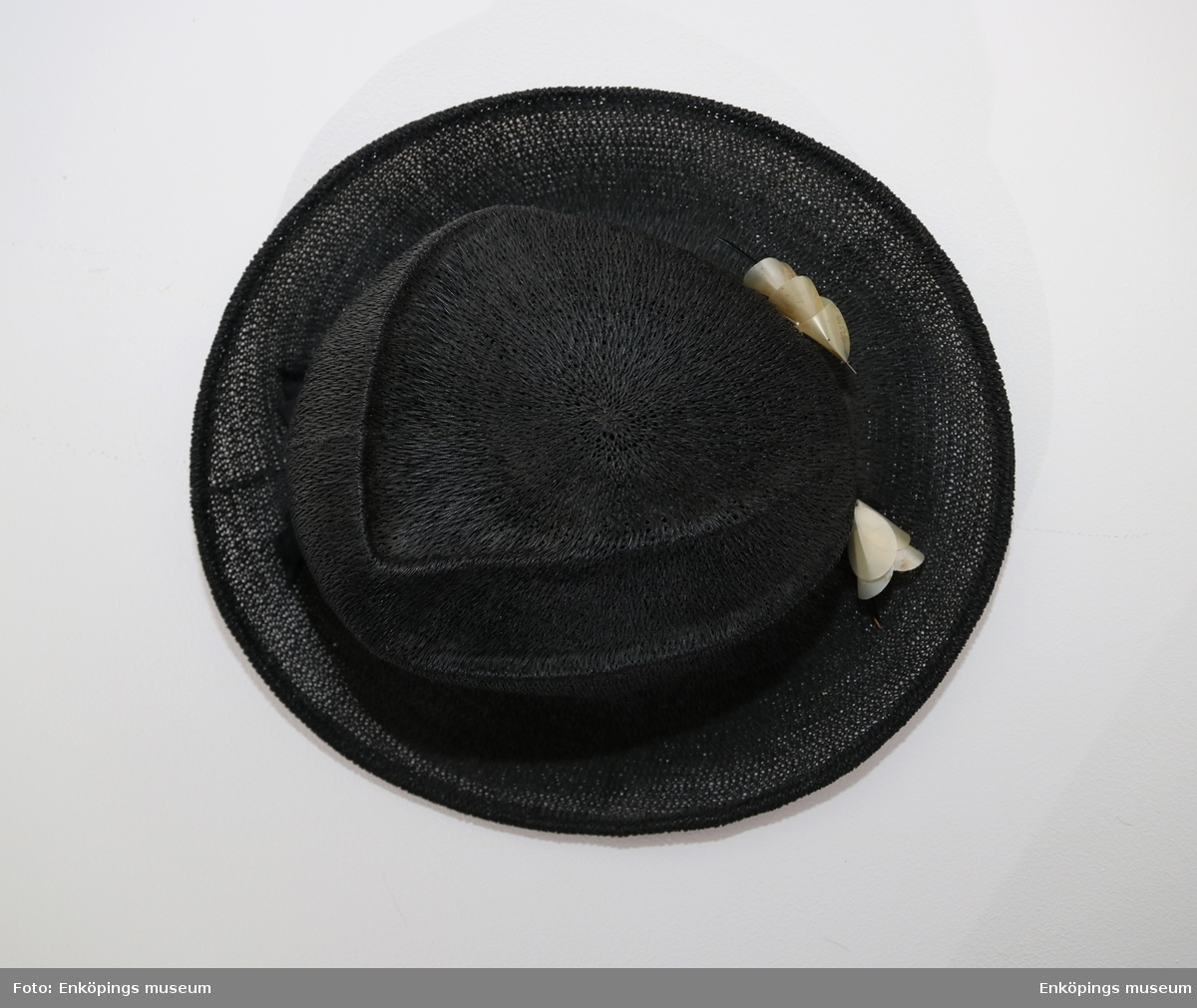 Svart hatt från Nya Mode & Tapisseriaffären, Enköping. Hatten har en rund modellerad kulle, svagt uppböjt brätte och svart ripsband runt kullen. Det finns en prydnad fram på ripsbandet, en ca 15 cm lång svart lackad trästicka med påträdda vita klockformiga prydnader.
