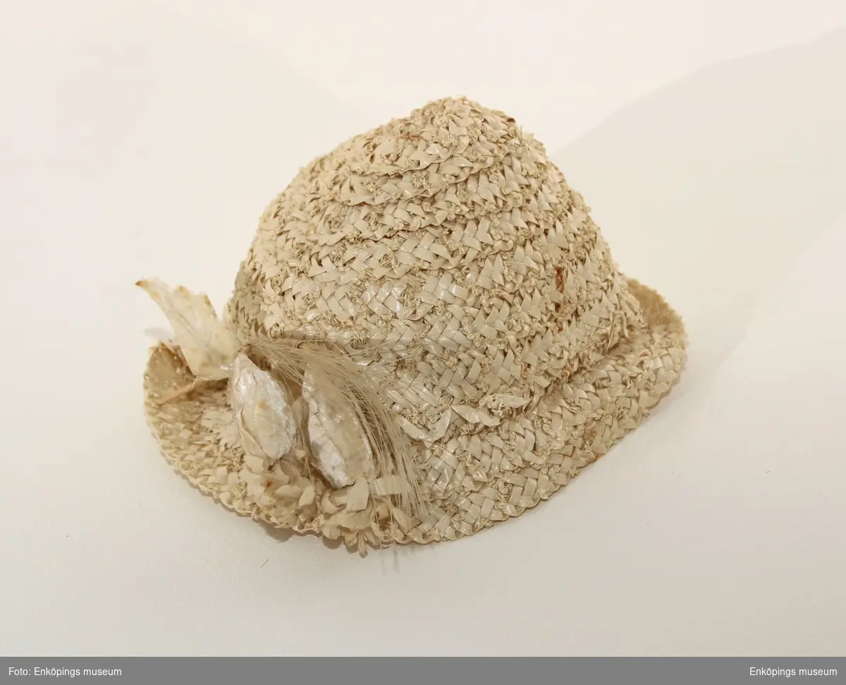 Krämvit hatt tillverkad av spiralsydda bastband. Brätte på halva rundeln. Dekor av två blomknoppslika former sydda av vit sammet, även stjälkar och blad av textil. Två " tofsar av tagel". Tillverkaren heter Ingrid Wik, Enköping, brukaren till denna hatt var Britt Lundevall.