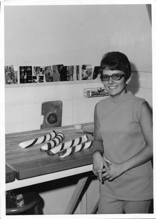 Okänd ung kvinna i ljus ärmlös dress med glasögon står lutad mot en arbetsbänk i ett polkagrisbageri. På bänken ligger fyra polkagrisar. På en list ovanför kakelplattor sitter flera vykort på rad.